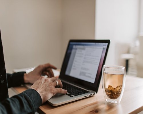 Close-up of hands typing on a laptop with a cup of tea nearby on a wooden desk